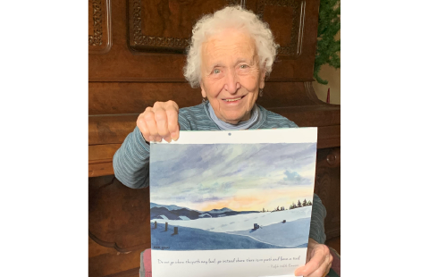 Barbara Favro, an older woman with short, white hair holds up the artwork "Norwegian Winter" from the 2023 IOD Calendar.