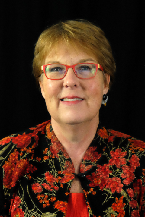 Denise Rozell is a woman with short reddish blond hair and red glasses wearing a black and red floral kimono style blouse