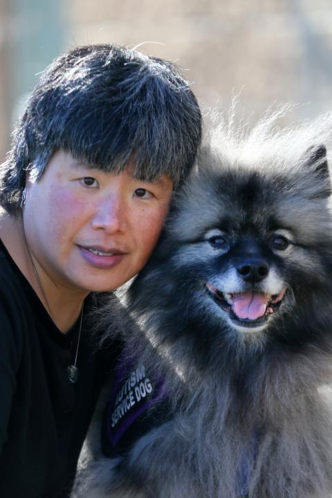 Amy Frechette is a woman with very short black and white hair posing with her service dog Judah, a keeshond who has fluffy gray and black fur