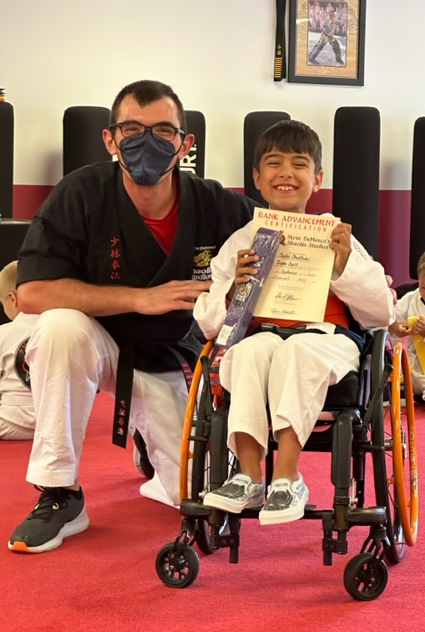 Bohdi smiles for the camera, holding his certificate of achievement and his new purple belt. His Sensei is down on one knee, next to Bohdi's wheelchair.