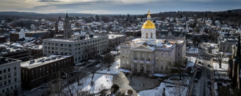 A wintertime aerial view of Concord, NH, featuring a large, stately building with a golden dome at its center. The building appears to be a government or historical structure, possibly a state capitol. Snow covers the surrounding ground, and the town's buildings stretch into the distance, with hills visible on the horizon.