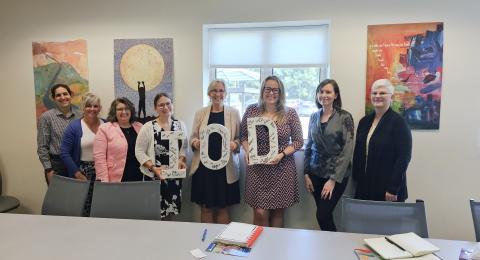 8 women posing in a conference room holding large letters with signatures on them that spell I O D