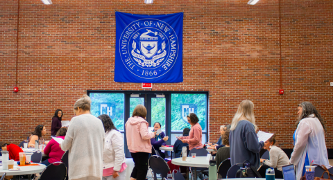 A large group of people are working and collaborating in one of the UNH event spaces, while a large blue banner hangs over the door with the words "The University of New Hampshire -1866 - 1893 1923 Science, Arts, Industry" with accompanying symbolism