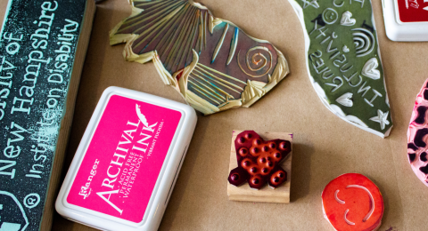 A handful of hand carved stamps and colorful ink pads are laid out on a table. One stamp has a heart made of brail dots, another looks like the shape of the state of NH with "Inclusion Matters" spelled out. Another stamp has University of New Hampshire Institute on Disability with the UNH shield logo on it.