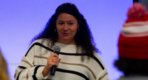 A woman with long curly hair stands comfortably speaking into a microphone to an attentive audience with a soft glowing screen behind her.