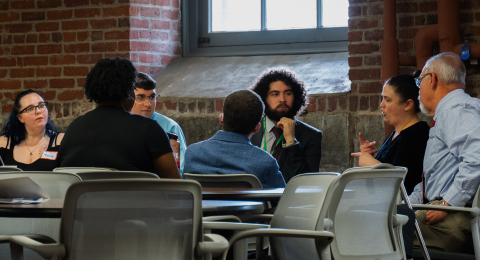 A diverse group of adult learners sits at a table paying attention while someone at the table signs.