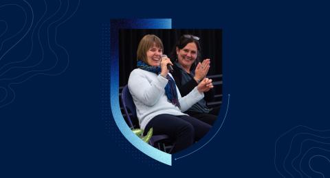 A swirling blue background with a shield cutout showing a photo of two smiling women sitting on a stage as one speaks into a microphone.