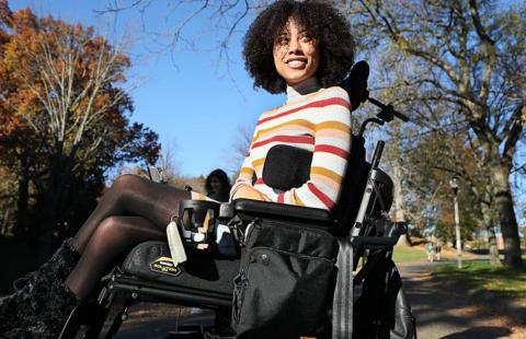 A woman of mixed race with shoulder length curly hair is wearing a striped turtleneck and sits in a wheelchair