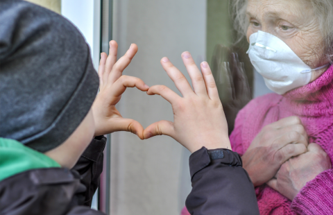 A child makes a heart symbol with his hands for his grandmother who is wearing a maks on the other side of a window.