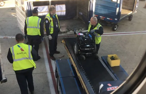 Airline workers on the tarmac loading a power wheelchair onto a conveyer belt leading to the plane's cargo hold.