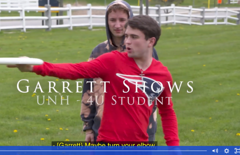 Garrett Shows wears a red Patriots long-sleeve t-shirt and shows a fellow student how to throw a frisbee on a lawn with a white fence.