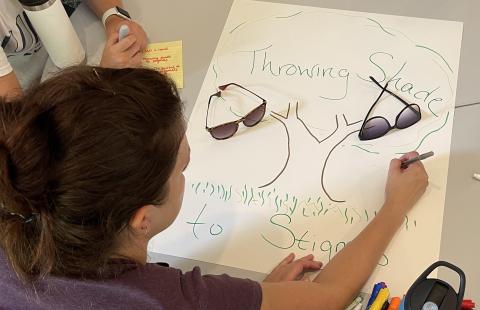 a couple of LEND trainees work on a poster of a tree with green grass below and the words Throwing Shade to Stigma written on it. There are two pairs of sunglasses resting on the poster as well.
