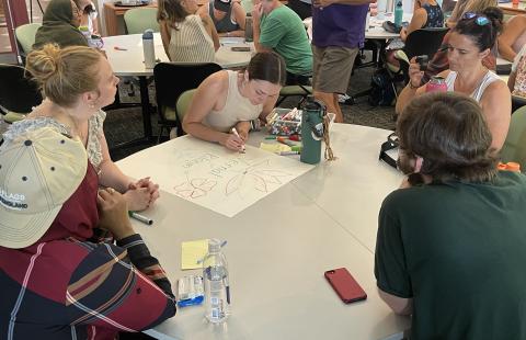 five LEND trainees gathered around a table, in a room full of other trainees working together at tables. One of the trainees at this table is drawing flowers on a poster that says Vernal Bloom