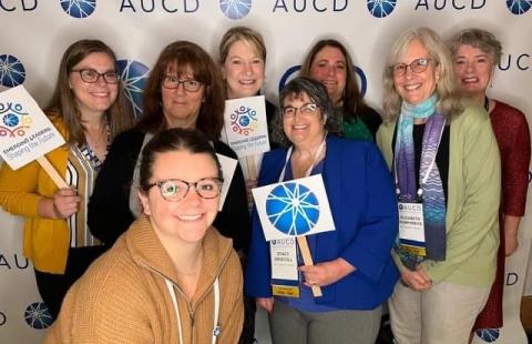 A group of women happily pose in front of an AUCD photo wall, some are hold little signs with the AUCD logo and Emerging Leaders: Shaping the Future