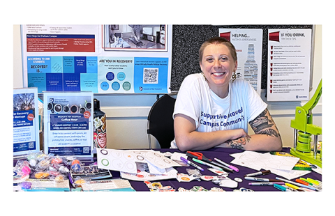 Heather Morris, a young white person with a buzzcut and a white t-shirt, smiles while hosting a resource table arrayed with flyers, candy, buttons, and stickers