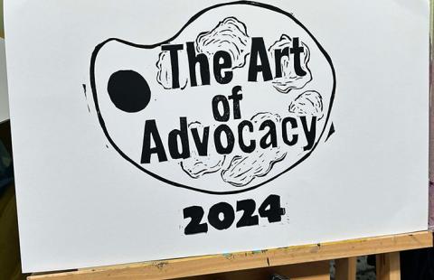 A wooden easel holds a white artboard with an black and white image of a paint palette and text reading The Art of Advocacy 2024