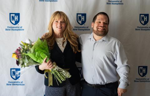Carrie Duran and Tim Houle pose together in front of a University of New Hampshire backdrop. Carrie, on the left, is holding a bouquet of flowers wrapped in green tissue paper and clear plastic. She has long blonde hair and is wearing a dark blazer and patterned pants. Tim, on the right, standing beside her with his arm around her shoulder, is wearing a light blue button-down shirt and dark pants. Both appear happy and celebratory.