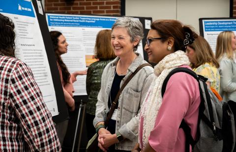 Photograph of a group of people attending a poster presentation event in an indoor setting, with multiple research posters displayed on easels. Attendees are engaged in discussions, with posters featuring titles related to health topics, and individuals wearing casual to business casual attire.