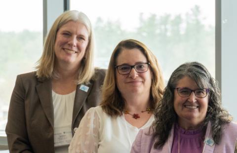 Photograph of three women standing indoors near a large window, suggesting a professional or conference setting.