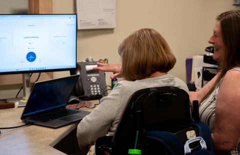 Two women, one in a wheelchair, sit at a desk happily working on setting up some new software on a computer.