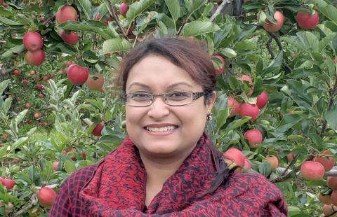 Tisha is a woman with glasses and brown hair pulled back, wearing a red/purple shirt and scarf standing in front of an apple tree