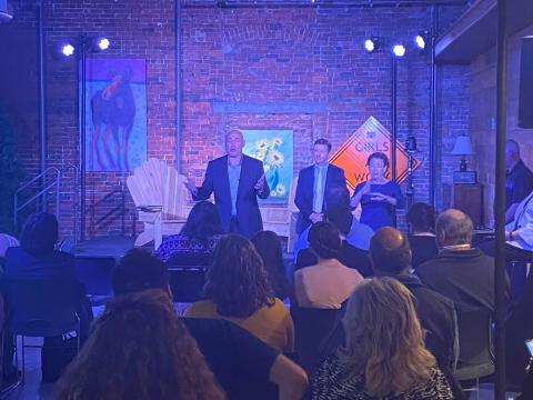 Dr. Tom Sherman, the moderator, and an ASL interpreter. There is an orange traffic sign that says, “Girls at Work” and a brick wall with art behind them. The audience is shown sitting in chairs in front of the stage. 