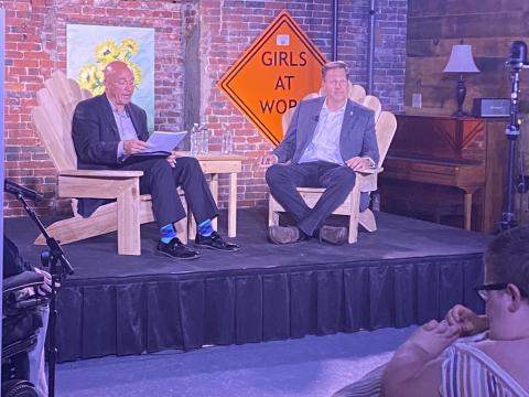 Governor Chris Sununu sits on stage with the moderator. There is an orange traffic sign that says, “Girls at Work” and a brick wall with art behind them.