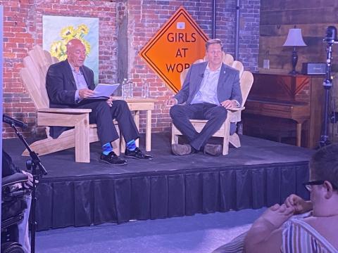 Governor Chris Sununu sits on stage with the moderator. There is an orange traffic sign that says, “Girls at Work” and a brick wall with art behind them. 