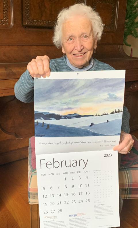 Barbara Favro, an older woman with short, white hair holds up the artwork "Norwegian Winter" from the 2023 IOD Calendar.