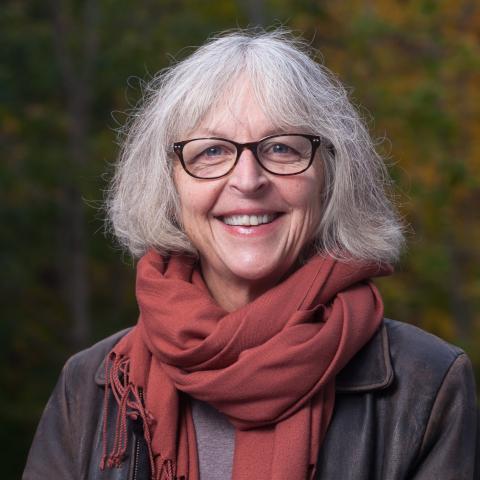 A middle-aged woman with shoulder-length gray hair, wearing glasses, a leather jacket, and an orange scarf.