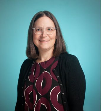 A middle-aged woman with short brown hair and glasses. She wears a shirt with maroon, white, and black swirls and a black cardigan.