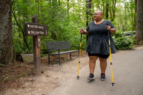 A disabled Black non-binary hiker uses trekking poles and looks at a wooden sign with “Nature House” and “Trillium Trail ♿” marked ahead. The shot is framed with the hiker walking toward the camera and the hiker sports a shaved head, glasses, a gray peplum shirt, black shorts, and black tennis shoes.