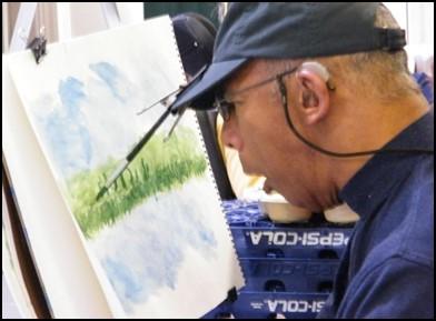 Tommy, an artist with Cerebral Palsy uses a paintbrush clipped to his baseball cap to paint a water color landscape
