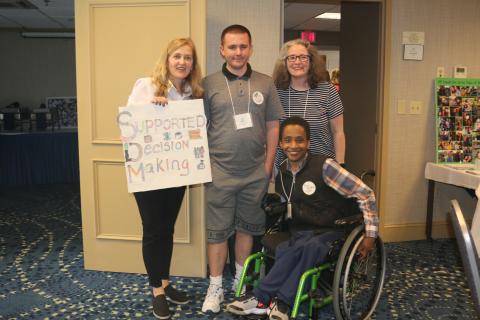 The 4 members of the Supported Decision Action Making Group pose together with one woman holding the sign. The group is made up of Meg, the sign holder and Michelle, two light skinned women, Nick, a light skinned man, and Eyob a medium-skinned man in a wheel chair.