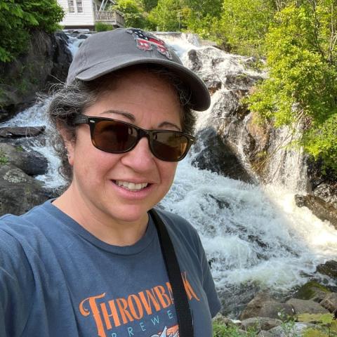 Deb Genthner is wearing a grey hat and brown sunglasses standing in front of a river flowing over rocks