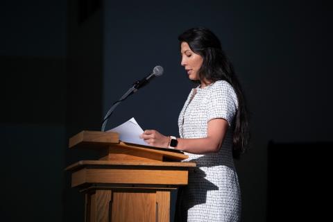 A woman with long wavy dark hair in light color tweed dress stands speaking at a podium, spotlighted in a dark room.