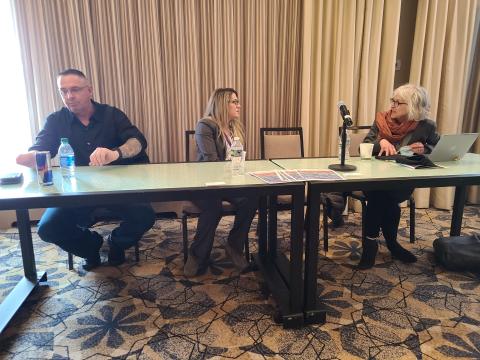 A man with short brown graying hair and glasses, a woman with long blond hair and glasses in a suit, and a woman with curly gray hair and glasses in a blazer sit at a presenter table together. The two women are chatting while the man is arranging his things.