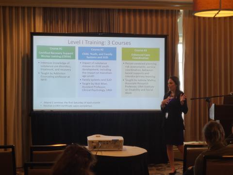 A woman with long brown hair, presents a powerpoint about Level I Training and the 3 courses involved to a conference room audience