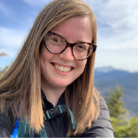 Katherine Russum is a woman with straight blond hair, wearing glasses and pictured outside with mountains and pine trees in the distance