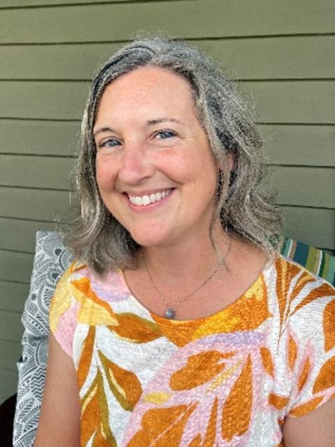 Tracey Miller is a woman with wavy midlength gray hair, wearing a colorful blouse sitting outside on a porch.