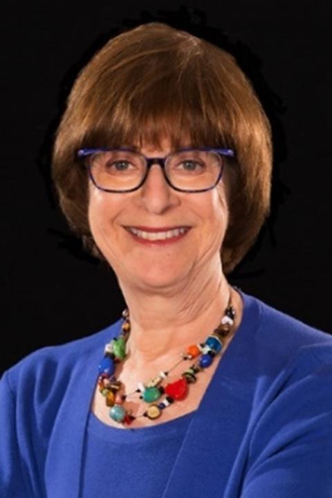 Elaine Katz is a woman with short auburn hair wearing glasses, a colorful necklace and bright blue sweater