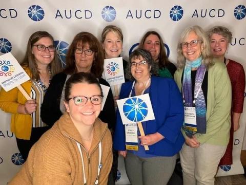 A group of women happily pose in front of an AUCD photo wall, some are hold little signs with the AUCD logo and Emerging Leaders: Shaping the Future
