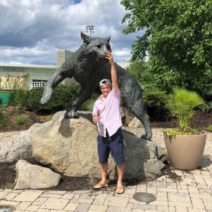 A young man in shorts, a button up polo, and a backwards ball cap,  reaches up above him to touch the nose of a big wildcat statue at UNH