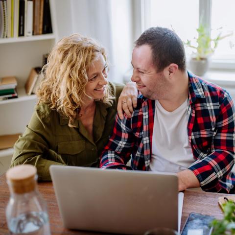 A man with down syndrome sits at a computer desk at home while a woman makes sure he's all set, they're smiling at each other.