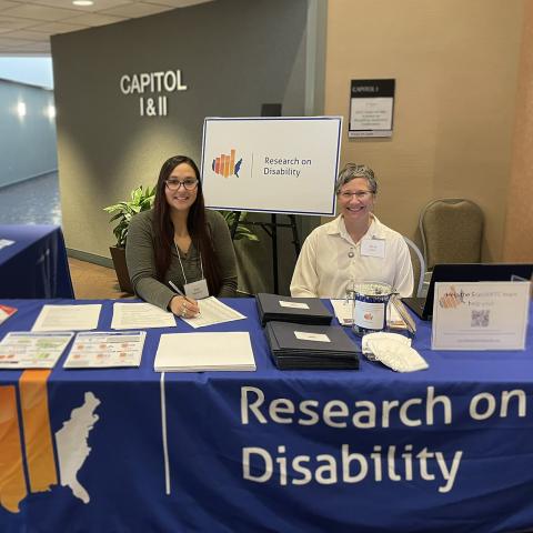 Two women sit at a conference registration check-in table for the Research on Disability State of the Science in DC