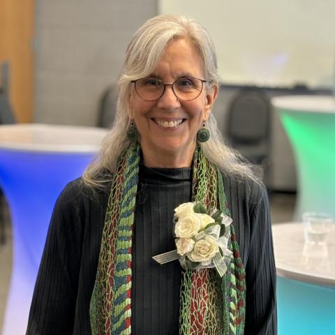 Betsy Humphreys is a happy smiling woman with long white hair, tortoise shell glasses, and a brightly colored scarf with a floral boutineer.