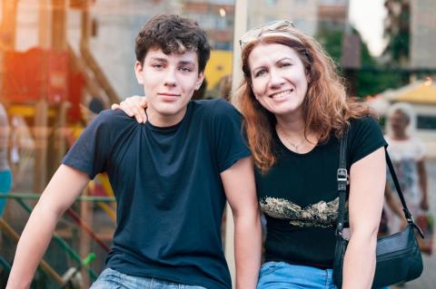 A mom puts her arm around her young adult son's shoulder. Both are wearing black tee-shirts and jeans.