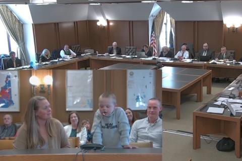 Screenshot of Meaghan Foster and her son Zander (a young boy with Downs syndrome) sitting in what looks to be a stateroom at a table, while Zander speaks into a microphone. This image is overlaying a wider shot of the committee itself at a large U shaped table arrangement..