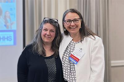 Michelle Lennon and Heidi Cloutier, two middle-aged white women, smile for a photo in a hotel conference room