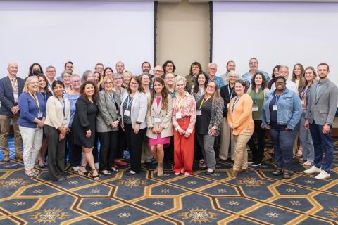 A group photo of about 40 diverse adults nicely dressed pose for a picture in a hotel conference space at the Human Services Workforce Development Symposium.